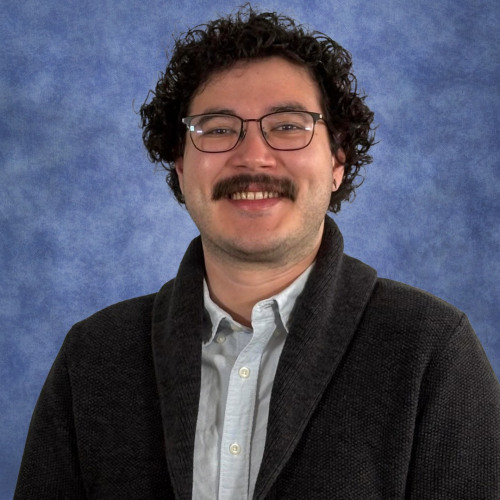Smiling young man with curly hair, glasses and a mustache wearing a button-down shirt and brown sweater.
