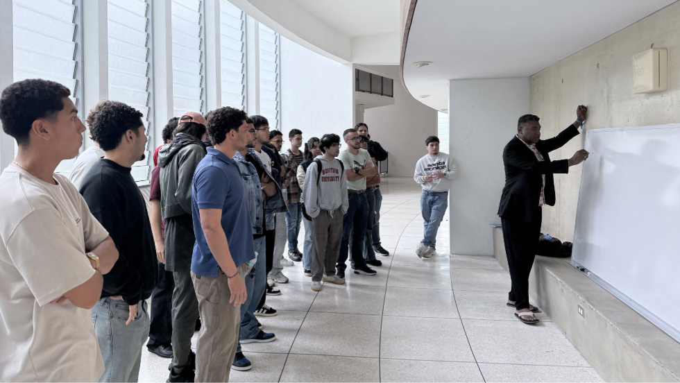 Instructor writes on a whiteboard while a group of students stand nearby watching in a bright indoor corridor.